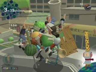 Screenshot from the video game We Love Katamari showing a colorful cluster of objects including fruits, furniture, and people, rolled into a large ball in a city environment, with a character in a red and white costume standing to the side.
