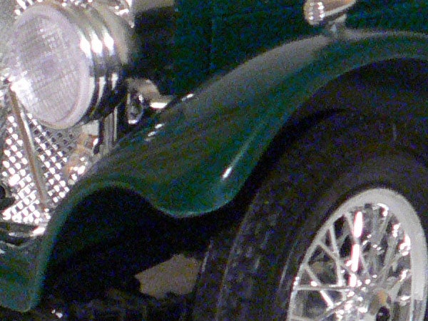 Close-up image of a green vintage car's front wheel and headlight, showcasing the detail and noise performance at high ISO captured by the Panasonic Lumix DMC-LX1.