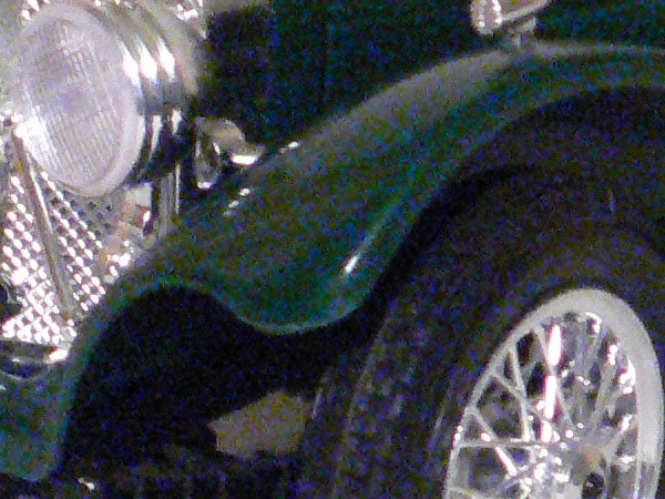 Close-up of a classic green car's front headlight, fender, and spoked wheel.