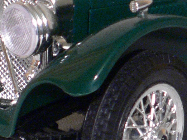 Close-up of a vintage green car's front left side showing details of the chrome headlight, wheel with spokes, and fender.