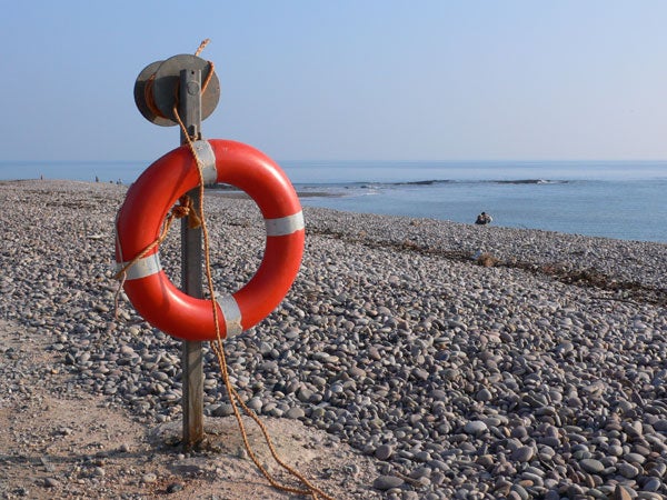 Red and white lifebuoy attached to a pole on a pebble-covered beach with calm sea and clear sky in the background, possibly taken with a Panasonic Lumix DMC-LX1.