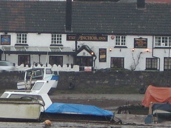 A photograph capturing the exterior of The Anchor Inn, a two-story building with signage and multiple windows, taken on an overcast day with visible boats covered with blue tarps in the foreground and a low tide exposing the ground beneath the boats.