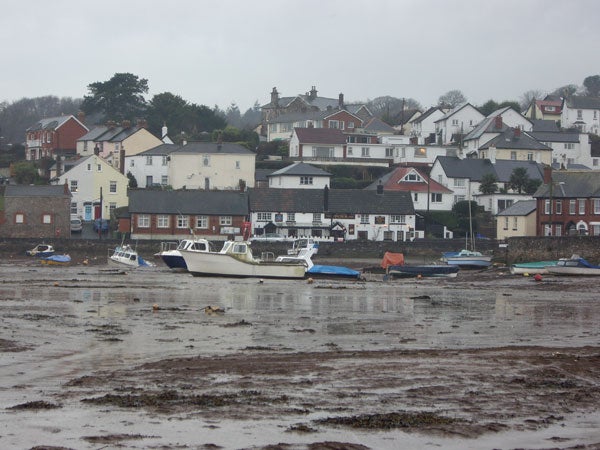 A coastal landscape depicting a low tide with boats resting on the seabed and a backdrop of residential houses on a cloudy day.A photograph capturing the exterior of The Anchor Inn, a two-story building with signage and multiple windows, taken on an overcast day with visible boats covered with blue tarps in the foreground and a low tide exposing the ground beneath the boats.