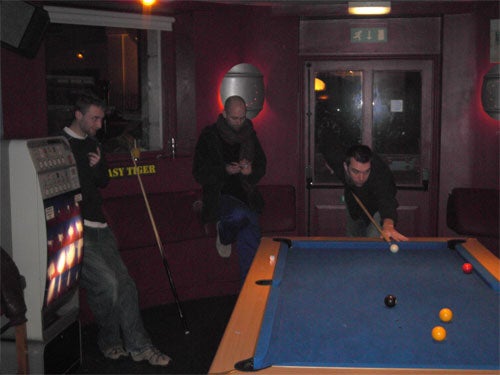 Three people in a dimly lit room with a pool table, one person is taking a shot while the other two watch.