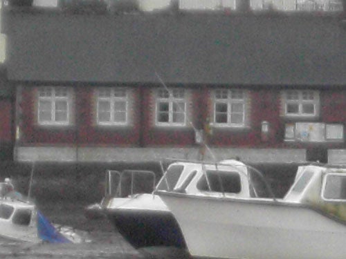 Photo taken with Acer CU-6530 camera showing boats moored near a waterfront with a red brick building in the background, image appears grainy, indicating low light performance.