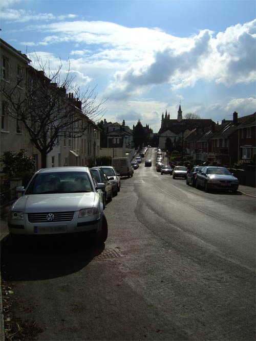 Image showing a street view with parked cars on the side, bright sky with clouds above, and buildings in the distance.
