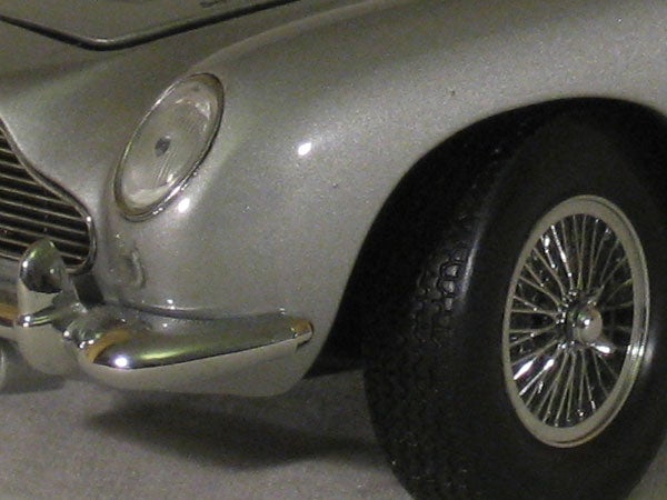 Close-up of a silver model car focusing on the front left side, highlighting the detail on the headlight, bumper, and spoked wheel.