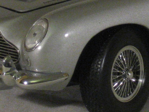 Close-up photo of the front section of a silver vintage car, highlighting the headlight, shiny chrome bumper, and spoked wheel.