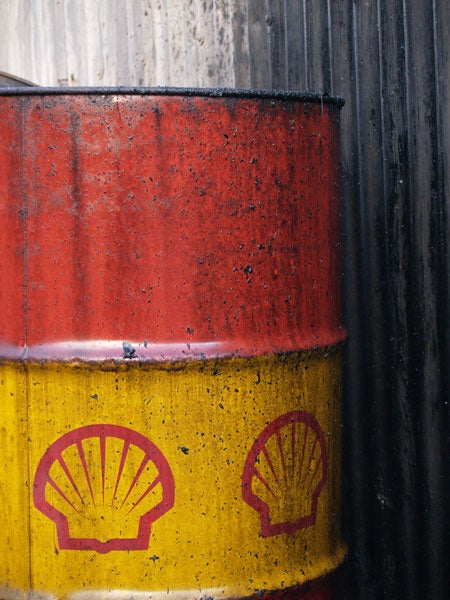 Close-up photo of a weathered oil barrel with red and yellow sections, each featuring a Shell logo, against a corrugated metal background.