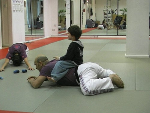 Martial arts training session with a child sitting on an adult's back who is lying face down on the mat, while another pair practices in the background.