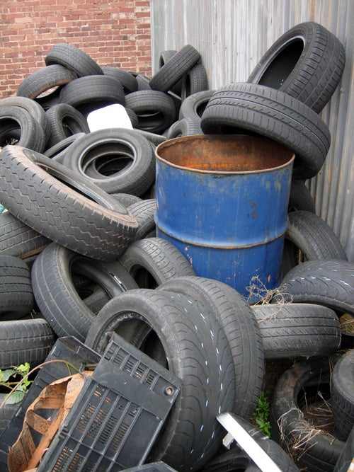 A pile of various discarded car tires heaped around a central, rusted blue barrel with a partially visible brick wall in the background.