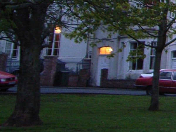 This image depicts a low-light outdoor scene taken during dusk or early evening, showing a street view with a tree in the foreground, a building with lit windows in the middle ground, and a red car to the left. The image appears to demonstrate the low-light imaging capability of a camera like the Ricoh GR Digital.
