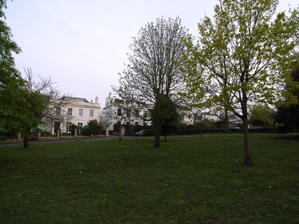Photograph taken by a Ricoh GR Digital camera capturing a park scene with green grass, trees in various stages of leafing, and a white building in the background under an overcast sky.