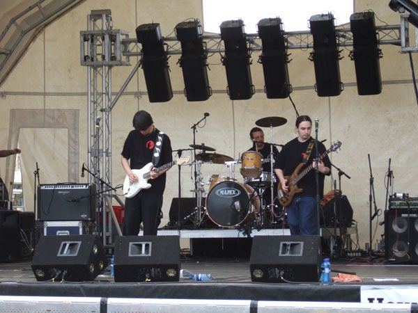A band performing live on stage with two guitarists and a drummer, captured by a Fujifilm Finepix F11 camera showing color accuracy and detail in an outdoor setting.
