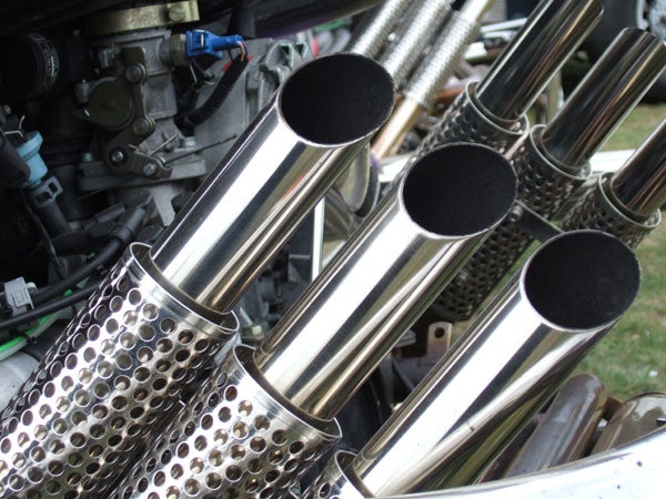 The image appears to be unrelated to the Fujifilm Finepix F11 or a product review. The picture shows a close-up of shiny, cylindrical metal exhaust pipes, possibly from a motorcycle or a car, with perforated heat shields around them and automotive parts in the background.