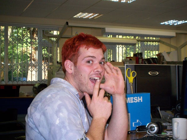 Man with dyed red hair sitting at an office desk, making a playful face and gesturing towards his cheek, with computer equipment and office supplies in the background.