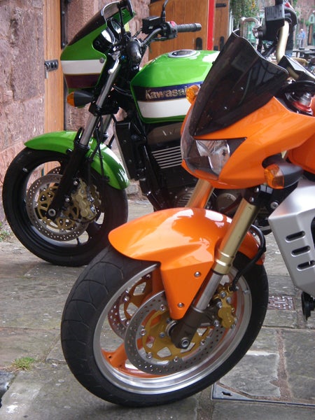 Photo of two parked motorcycles, one orange and one green, with a close-up view of the orange motorcycle's front wheel and brakes.