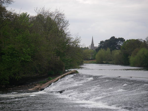 A scenic river view with a small waterfall, lush green trees along the banks, and a church spire in the distance under a cloudy sky.