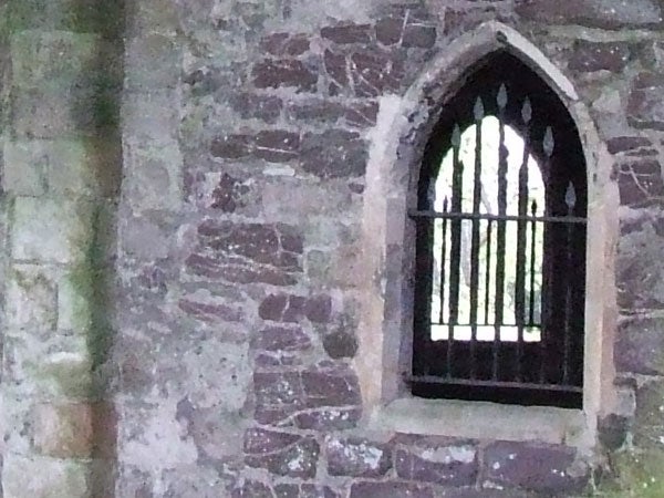Photograph taken with a Fujifilm FinePix S5600, featuring an old stone wall with a Gothic style arched window with iron bars.