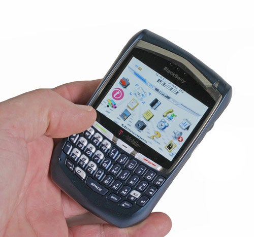 A person holding a T-Mobile BlackBerry 8700 smartphone with its screen turned on displaying icons and applications. The phone has a full QWERTY keyboard and a T-Mobile logo is visible on the screen.