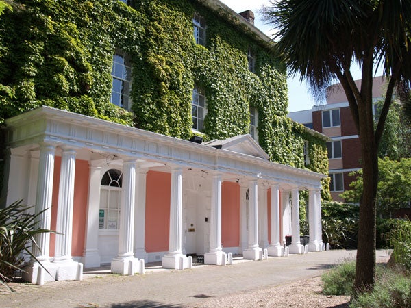A photograph showcasing a two-story building covered in verdant ivy, featuring a classical facade with pink columns and a series of white entry doors, taken during the day under clear blue skies.