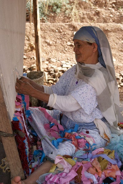 Elderly woman weaving on a loom outdoors.