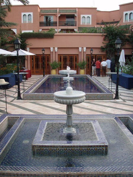 A photograph showing an elegant courtyard with a rectangular pool and a smaller square fountain in the foreground, surrounded by mosaic tiles. In the background, there is a pink building with a balcony, several guests, and restaurant-style outdoor umbrellas.