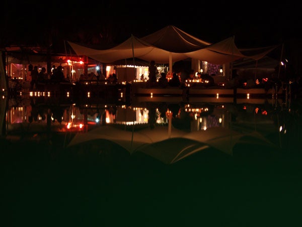 Nighttime scene showing a poolside bar with illuminated tents and reflection on the water, captured in low-light conditions indicating the camera's performance in such environments.