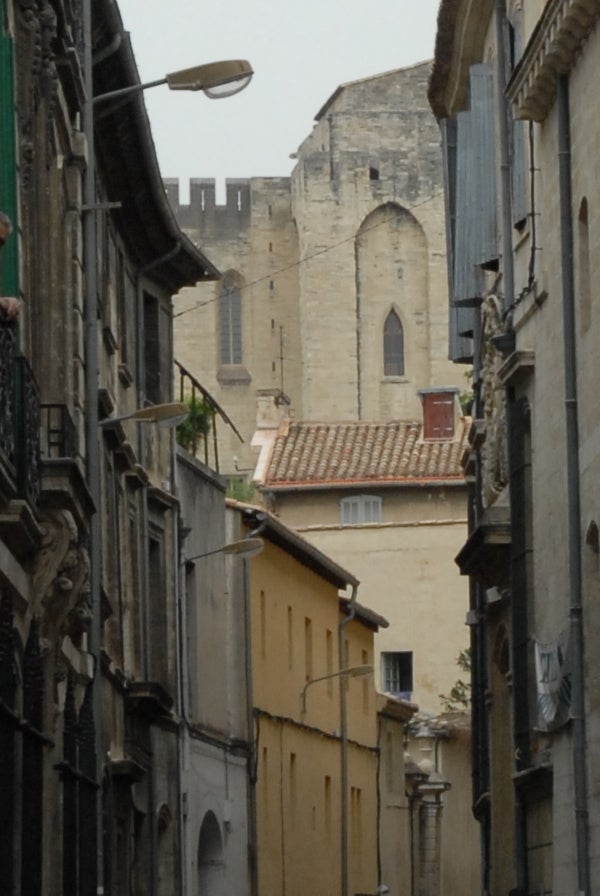 A narrow European street flanked by tall, weathered buildings with balconies, leading to a view of an ancient stone church with battlements under an overcast sky, likely taken with a Nikon D200 digital SLR camera.