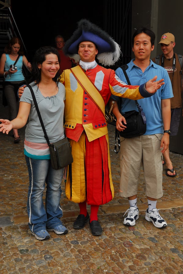 Three people posing for a photo with a man dressed in a colorful traditional European outfit including a tricorne hat. The individuals are standing on a cobblestone surface with a crowd visible in the background.