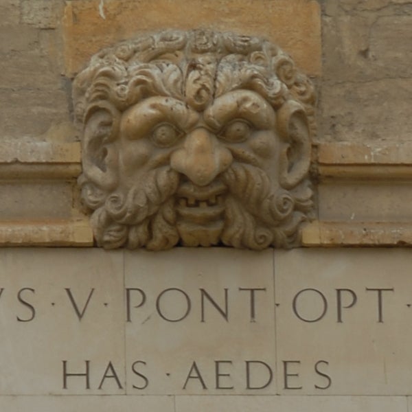 Stone carving of a bearded man's face on a building facade with inscribed Latin text below.