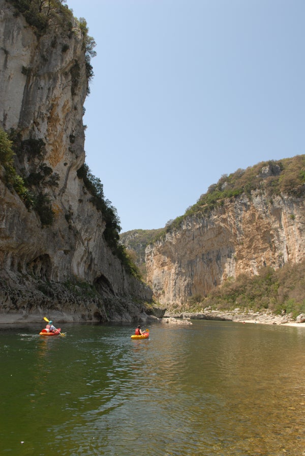 Two people kayaking in a river with steep cliff walls on either side under a clear blue sky.