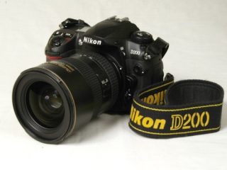 Nikon D200 digital SLR camera with a zoom lens attached and a camera strap beside it, displayed against a plain background.