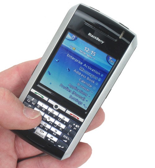 A hand holding a BlackBerry 7130g smartphone with a blue screen showing options for enterprise activation, address book, calendar, messages, and browser. The phone has a full QWERTY keyboard and is silver and black in color.