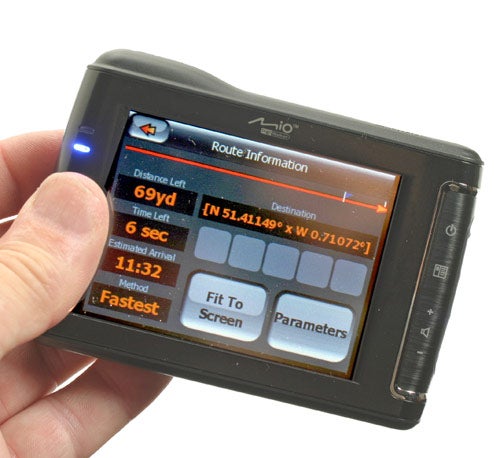 Hand holding a Mio DigiWalker C710 GPS navigator displaying route information with options for navigational parameters on the screen.