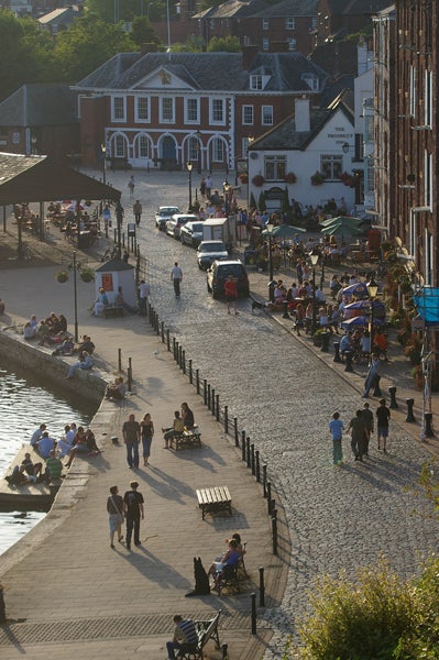 Crowded riverside walkway with people sitting at outdoor tables and walking along a cobblestone street, with historic buildings and parked cars in the evening sun.