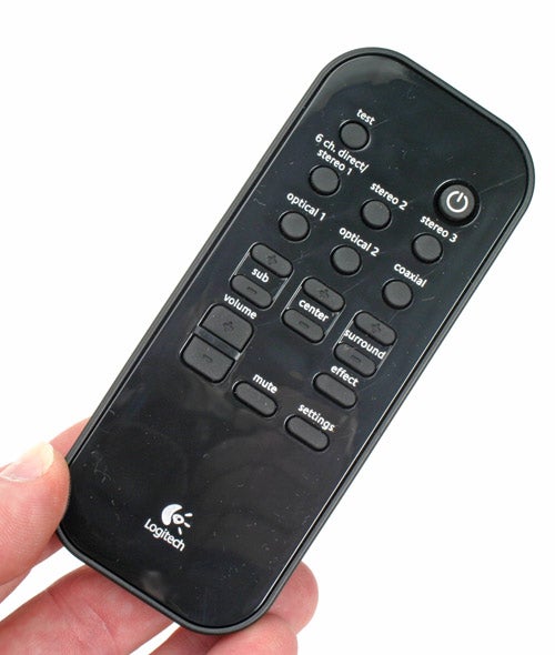 A person holding a black Logitech remote control with buttons for test, input selection, sound effects, volume, and system power. The Logitech logo is visible at the bottom. The remote displays signs of use and fingerprints.