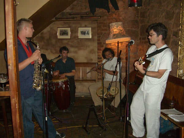 Four musicians performing in a dimly lit, cozy pub with stone walls, two playing saxophone and electric guitar at the front and two playing percussion and keyboard in the background.