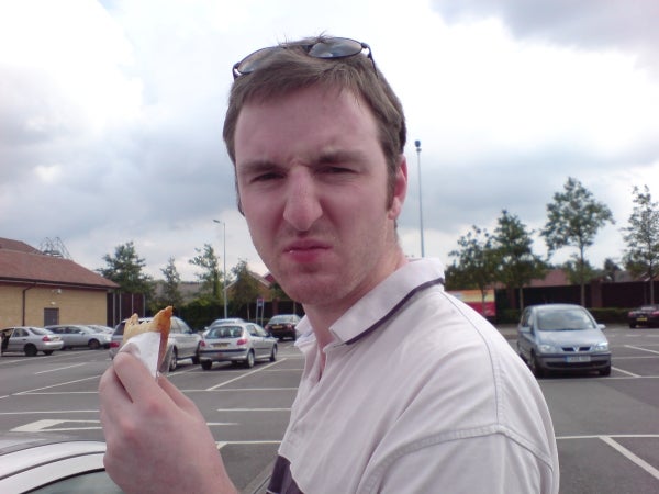 A man with a skeptical expression eating a sandwich in a parking lot.