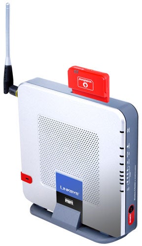 Linksys Wireless-G Router for 3G/UMTS Broadband standing vertically on a surface with an antenna on the left side, status LEDs on the front panel, and a red 3G dongle inserted on the top.