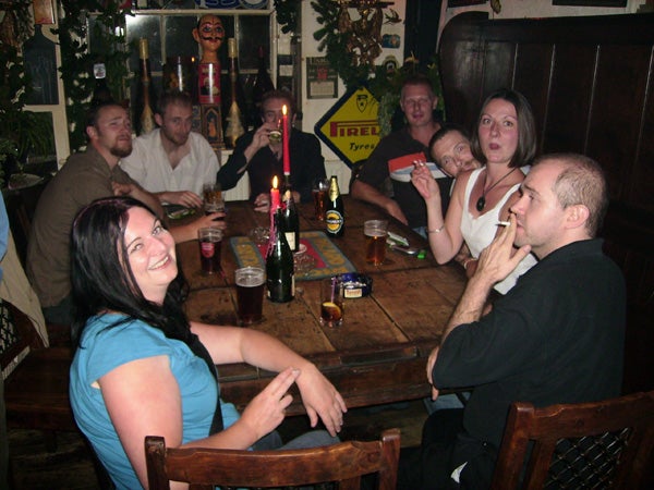 A group of people sitting around a wooden table in a dimly lit pub with drinks, interacting and smiling, possibly taken with a low-light capable camera like the Olympus FE-150.
