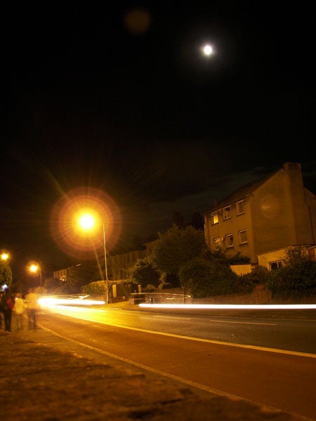 Long exposure night photograph taken with Kodak EasyShare P712, showing streaks of light from moving vehicles, a brightly illuminated street lamp, a row of houses, and the moon in the sky.