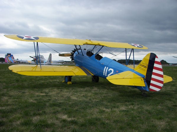 The image displays a biplane with yellow wings and a blue fuselage sitting on a grassy field. The tail is striped in red and white, and the number 112 is visible on the side. Airplanes can be seen in the background under an overcast sky. This image appears unrelated to the Kodak EasyShare P712 product or its review.