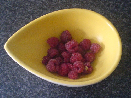 A yellow, triangular bowl containing fresh raspberries on a kitchen countertop.