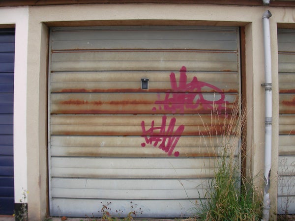 Rusted metal garage door with purple graffiti and overgrown grass in the foreground.