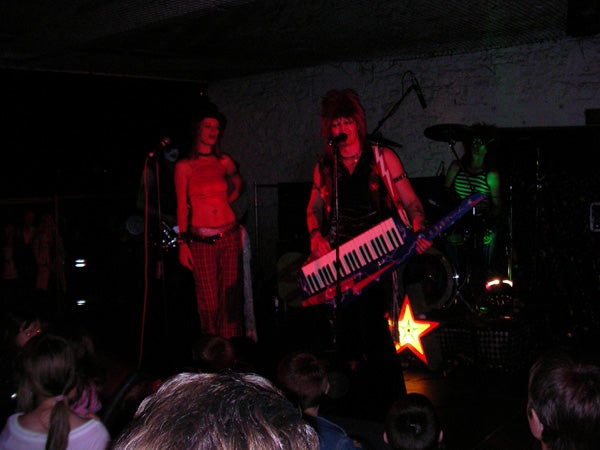 Band performing on stage, with a member playing a keytar while another stands near the microphone, in a dimly lit venue with an audience in the foreground.