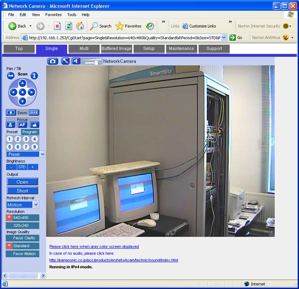 A screenshot of a Panasonic BB-HCM381 IP Camera's interface on a computer monitor, showing a live feed from the camera with a view of a tech room with two computer monitors, a large server unit, and various cables.