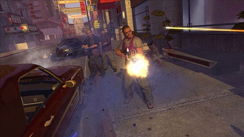 A screenshot from the video game Saints Row showing a character in the middle of a street shootout with police, firing a gun with visible muzzle flash, surrounded by chaos, with damaged vehicles and urban shops in the background.