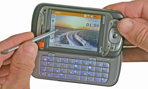 Hands holding an Orange SPV M3100 smartphone with a stylus interacting with its touchscreen, displaying the start screen with a QWERTY keyboard extended.