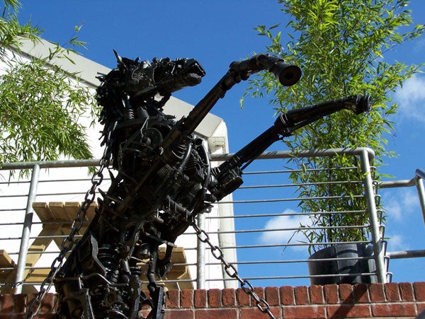 A metal sculpture of a violin player with mechanical details, displayed outdoors near green shrubbery and a brick wall, with a blue sky in the background.